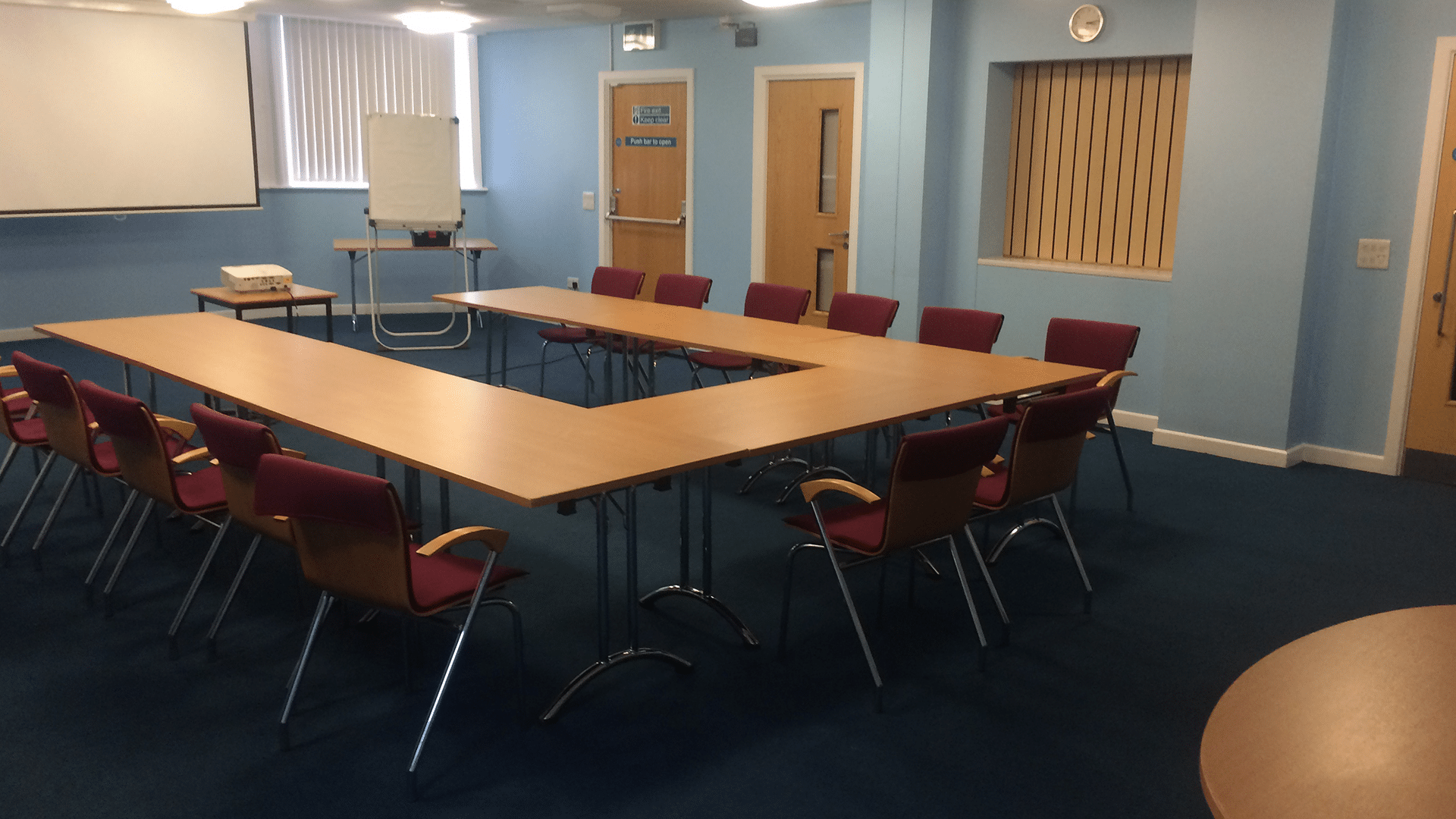 Interior of the Beacon Room, the table is set up in a U-shape formation with red chairs and a projector.